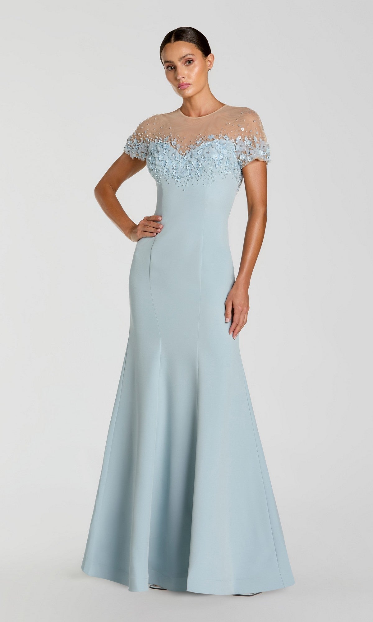 A woman wears the Mac Duggal 11867 short sleeve long formal dress in light blue, featuring sheer sleeves and floral embellished bodice, standing against a plain background.